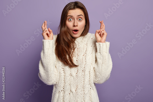 Superstitious shocked woman crosses fingers, hopes to make good deal, makes wish, has bated breath, dressed in loose white sweater, isolated over purple background. People and expectation concept