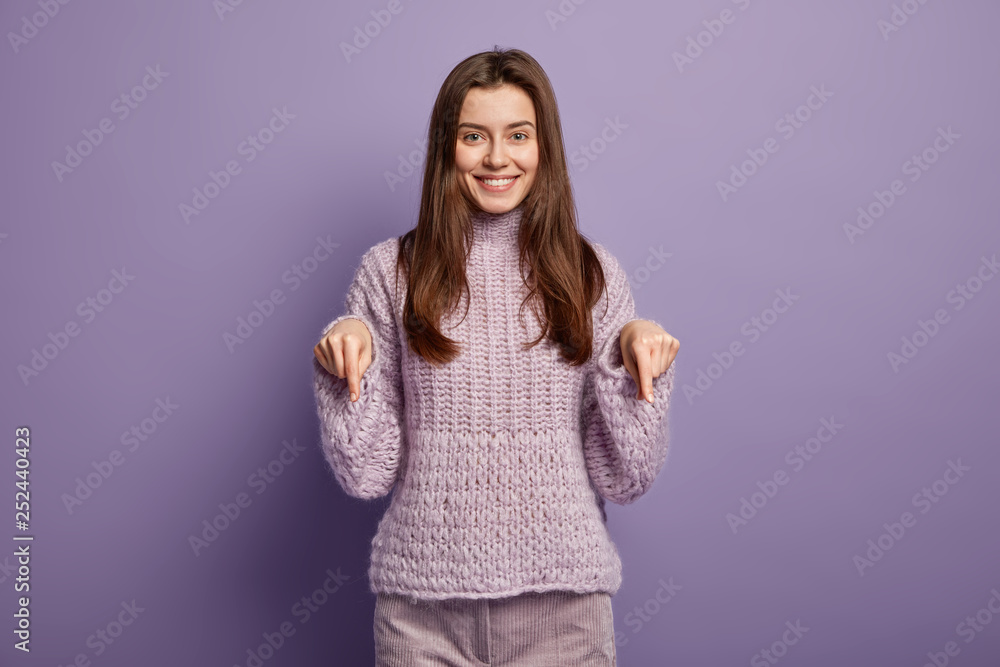 Gorgeous smiling young woman points down on floor, wears knitted sweater and corduroys trousers, advertises new shoes she bought in shop, isolated over purple backgroud. Advertisement concept Gorgeous smiling young woman points down on floor, wears knitted sweater and corduroys trousers, advertises new shoes she bought in shop, isolated over purple backgroud. Advertisement concept