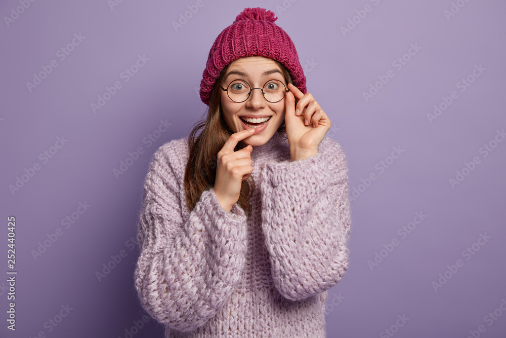 Horizontal shot of optimistic glad woman keeps one hand near mouth, other on rim of glasses, smiles positively at camera, feels energized and upbeat has fun over purple background wears winter clothes