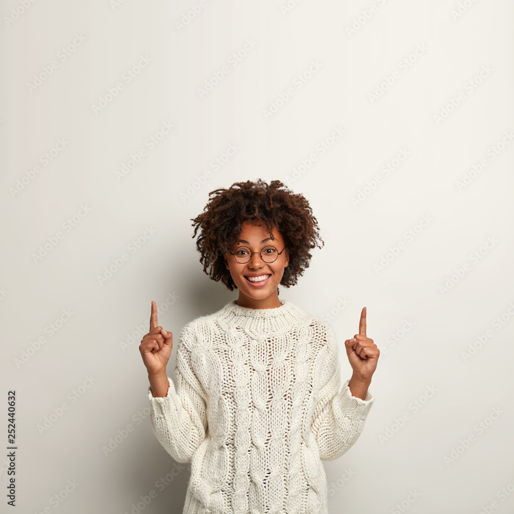 Good looking optimistic dark skinned girl shows item upwards, points with both fore fingers, wears spectacles, feels joyful isolated over white background. Curly African American woman sells something Good looking optimistic dark skinned girl shows item upwards, points with both fore fingers, wears spectacles, feels joyful isolated over white background. Curly African American woman sells something