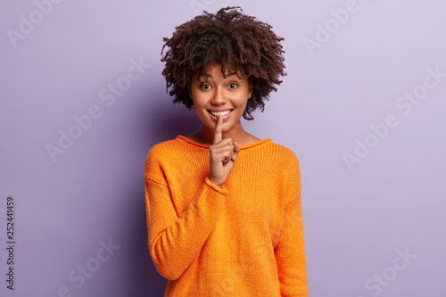 Positive woman with cheerful expression makes hush gesture  asks not spread rumors  dressed in orange clothing  isolated on purple background  orders keep silent. Shut your mouth. Let us make surprise