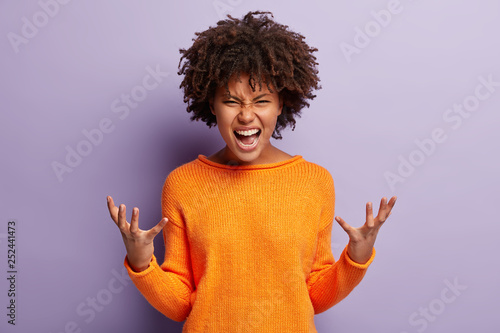 Foto Angry annoyed female gestures with annoyance, screams loudly, expresses irritation, wears casual orange jumper, reproaches partner in betrayal, isolated over purple studio wall