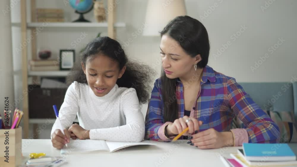 Medium shot of asian happy woman tutor teaching and helping her black african american girl student at home. African child and her mixed race school female teacher working together on wooden table Medium shot of asian happy woman tutor teaching and helping her black african american girl student at home. African child and her mixed race school female teacher working together on wooden table