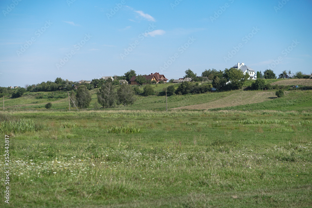 Countryside view near a small river and big hills with houses. Beautiful landscape with trees and meadow.