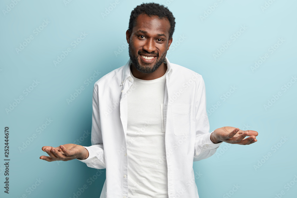 Horizontal view of happy careless guy, spreads hands with clueless facial expression, has no idea, makes choice between two pleasant things, wears white stylish outfit, models over blue background. Horizontal view of happy careless guy, spreads hands with clueless facial expression, has no idea, makes choice between two pleasant things, wears white stylish outfit, models over blue background.