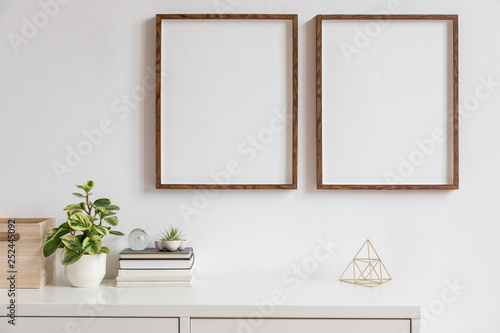 Stylish white home decor of interior with two brown wooden mock up photo frames with books, beautiful plant in stylish pot, gold pyramid and home accessories. Minimalistic scandinavian room.