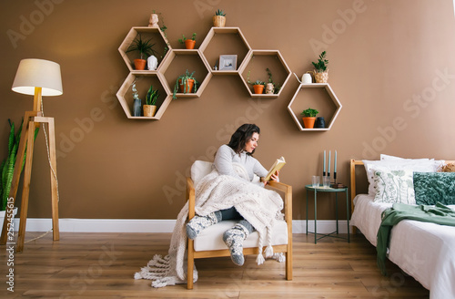 Cute woman enjoying reading a book sitting on armchair in apartment
