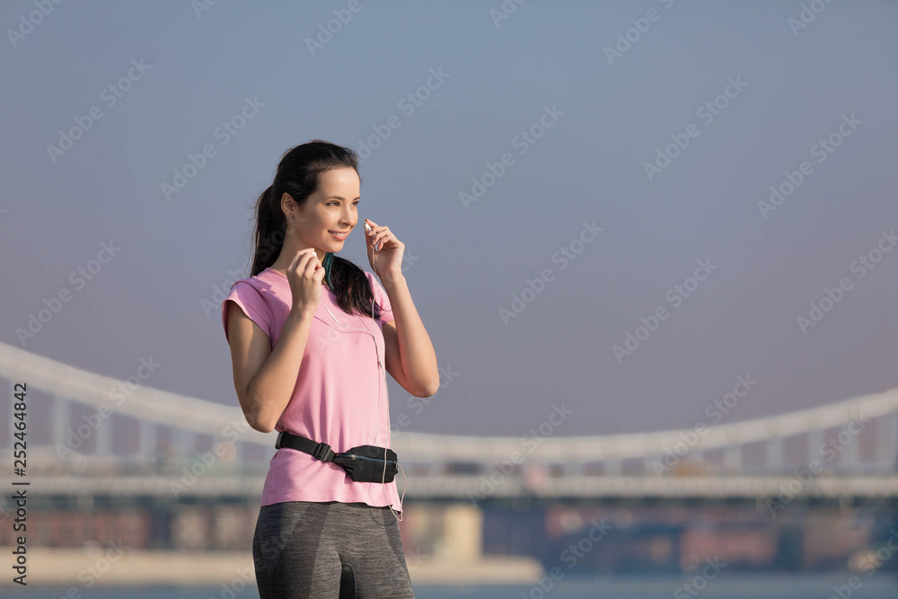 Attractive caucasian girl in a pink t-shirt and gray sports trousers listens to music at sunset on the street. On a background Krymsky Bridge