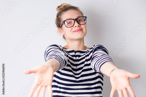 Beautiful woman happy and excited expressing pulling hands to hug. Smiling joyful blonde girl wearing glasses and striped dress on white