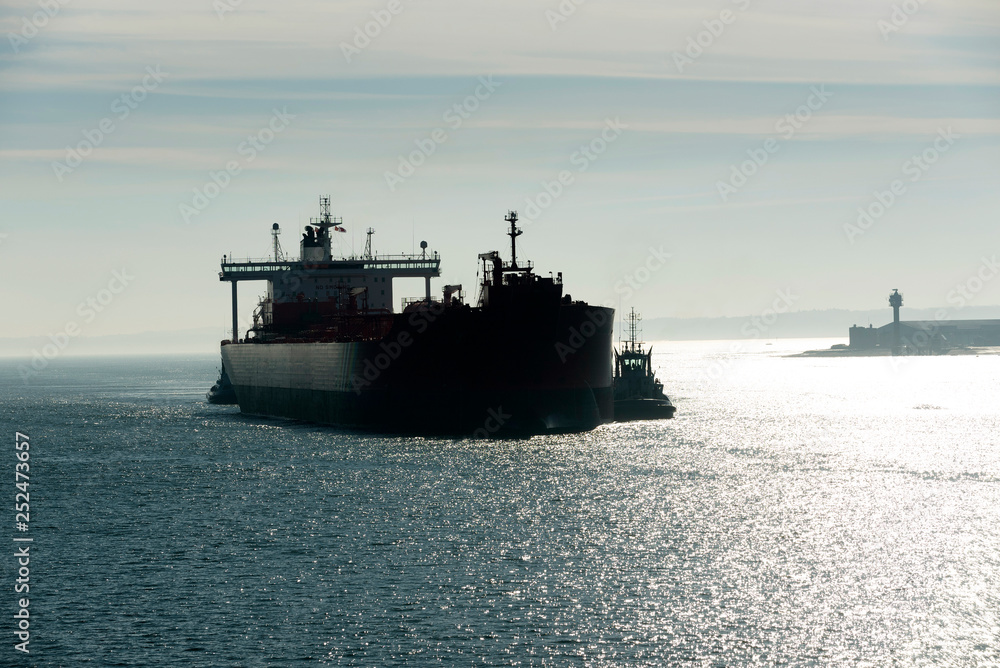 Southampton Water, Hampshire, England, UK. February 2019. Early morning against the light shot of a tug in attendance moving a broad beam oil tanker onto a berth at Fawley refinery.