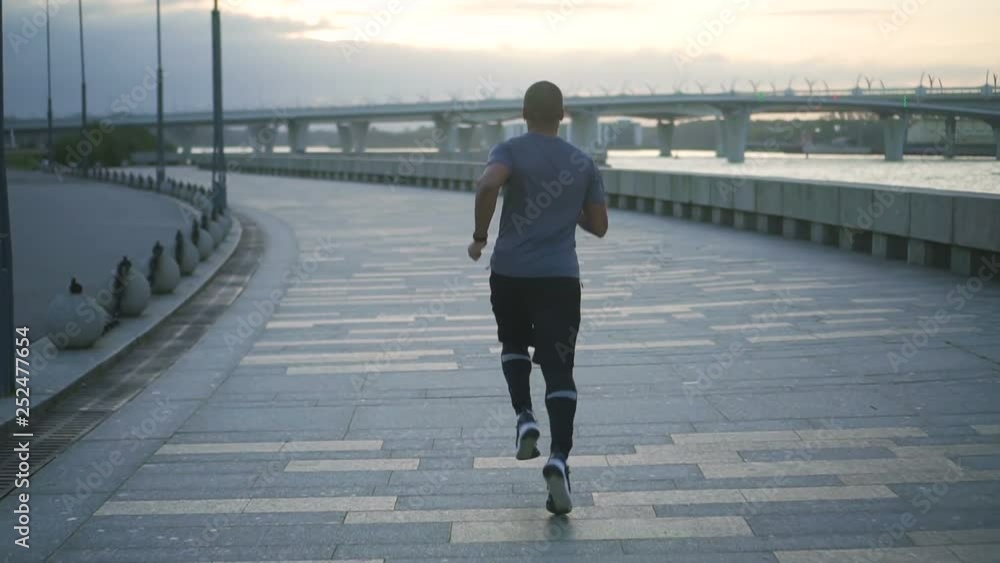 Young healthy black man training and building his body doing fithness and exercise on sidewalk in slow motion. Behind view of african american active person male jogging and running early in the Young healthy black man training and building his body doing fithness and exercise on sidewalk in slow motion. Behind view of african american active person male jogging and running early in the