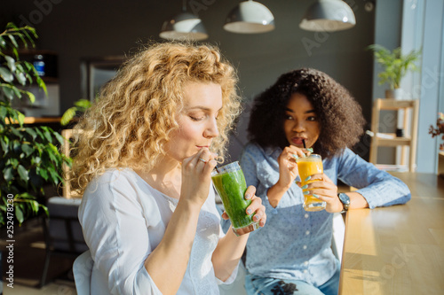 Cheerful multi-ethnic curly women having fun at fast food restaurant. Two female friends with healthy cold drink glass on table at cafe or restaurant interior.