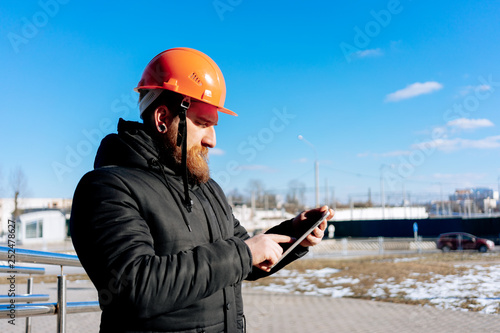 Pretty man - specialist civil engineer at orange hard hat is controls his project (outdoors). Blue sky and modern architecture (buildings) on background. Sunny day. photo