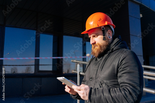 Pretty man - specialist civil engineer at orange hard hat is controls his project (outdoors). Blue sky and modern architecture (buildings) on background. Sunny day.