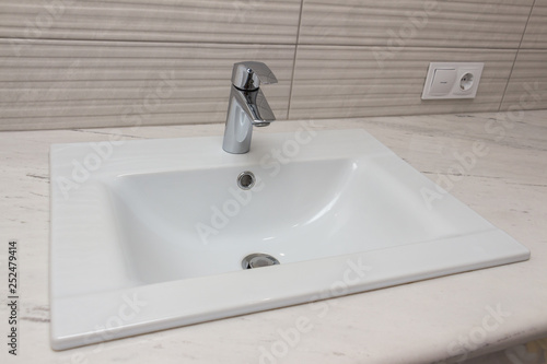 Ceramic white washbasin with stainless faucet mounted on a marble worktop against a background of gray large wall ceramic tiles. Bathroom interior element, close-up