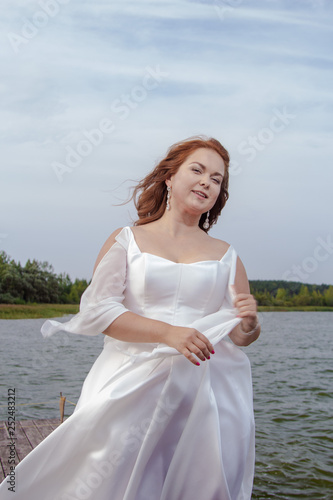 Red-haired bride in a white dress posing on a quay near a lake photo