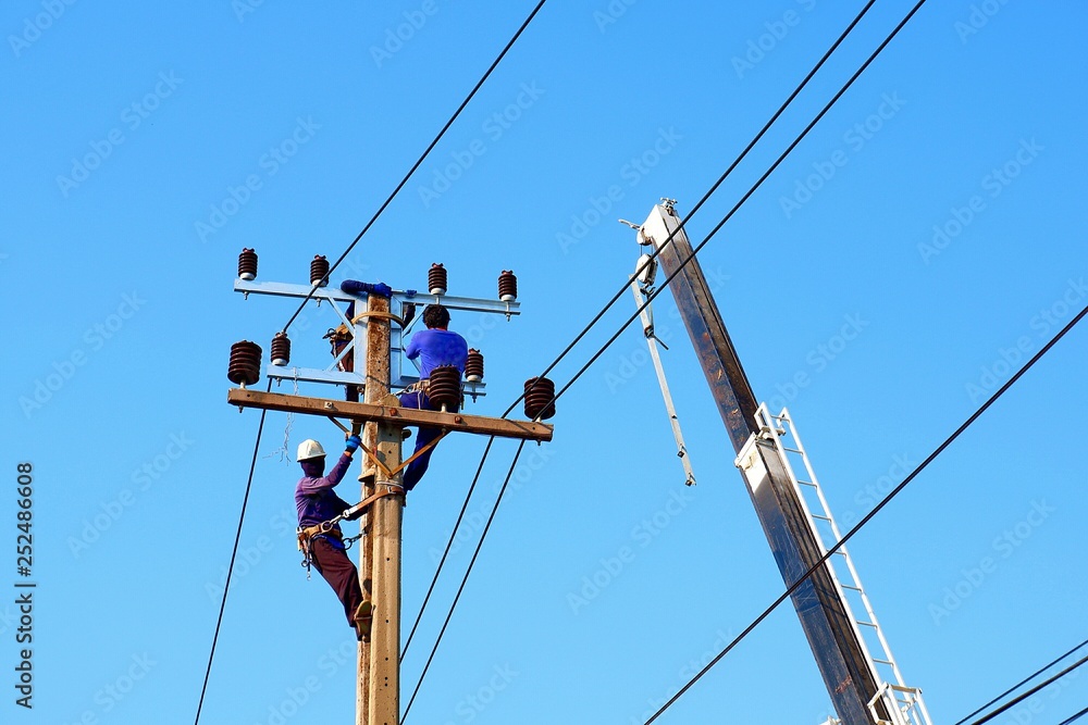 Low angle view of electricians team with crane are working to install electrical transmission equipment on power pole against blue clear sky background Low angle view of electricians team with crane are working to install electrical transmission equipment on power pole against blue clear sky background