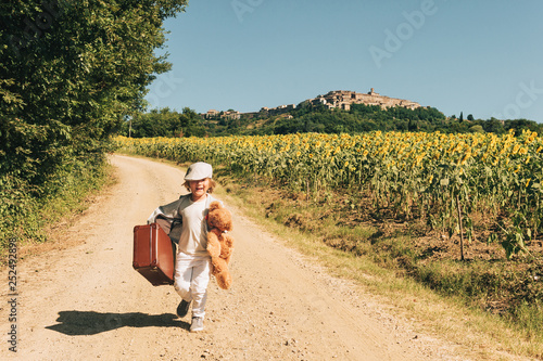 Outdoor portrait of a funny little boy walking down the road in countryside, holding old small suitcase and teddy bear toy. Image taken in Tuscany, Italy