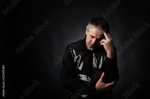 Anxious handsome male with grey hair in expensive fashion shirt sitting in dark, holding hand on head, thinking about problems. Copy space on left.