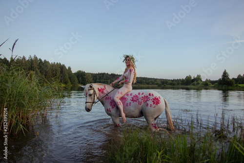 Blonde girl in white and red bodypain bathes with a horse in the lake at sunset. photo