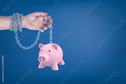 Close-up of man's hand with metal chain around wrist holding piggy bank hanging on the other end of chain. photo