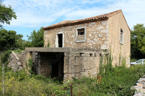 Abandoned traditional stone house with destroyed windows and doors with large basement in front covered with terrace and surrounded with high grass and other plants on cloudy blue sky background