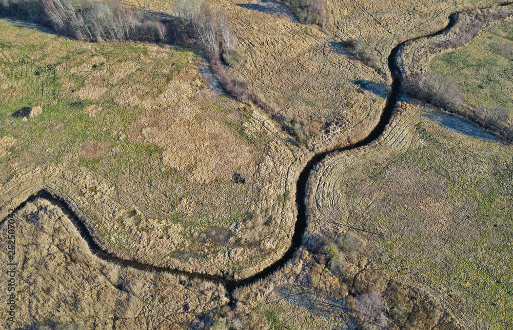 Aerial view on river between trees and yellow meadows during winter.