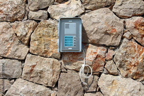 Brand new door entry intercom system mounted on traditional stone wall with hanging electrical wires waiting to be connected on warm sunny day