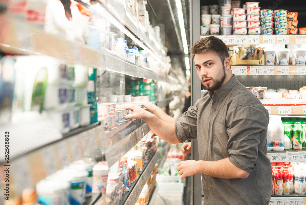 Portrait of amazed man standing near the refrigerator in the milk department of the supermarket and looking at the camera. Buyer chooses yogurt in a grocery store, looks at the camera with amazement.