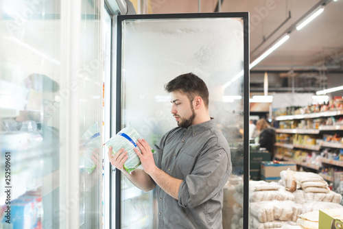 Buyer breaks the refrigerator door in the supermarket, holds the package in his hands and looks at the label. Man chooses frozen food in a supermarket fridge. Choice of frozen foods in the supermarket