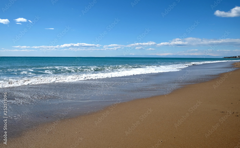 Beautiful landscape with sea, surf with waves with foam runs to long coast with empty sandy beach and high mountains with snow caps on skyline in haze on sunny summer day