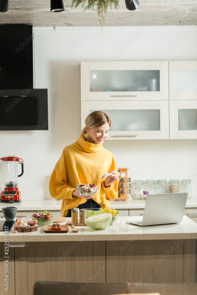 Happy young lady looking at the screen of laptop while cooking