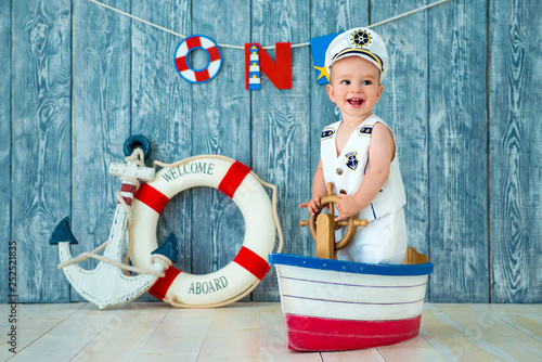 Photoshoot for a boy of one year. Little sea captain, sailor on toy ship with steering wheel. Sea anchor and lifebuoy on gray wooden background photo