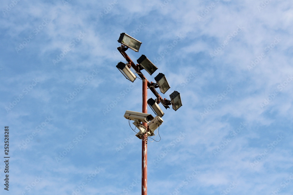 Obraz premium High metal pole with multiple light reflectors and security camera connected with electrical wires on cloudy blue sky background