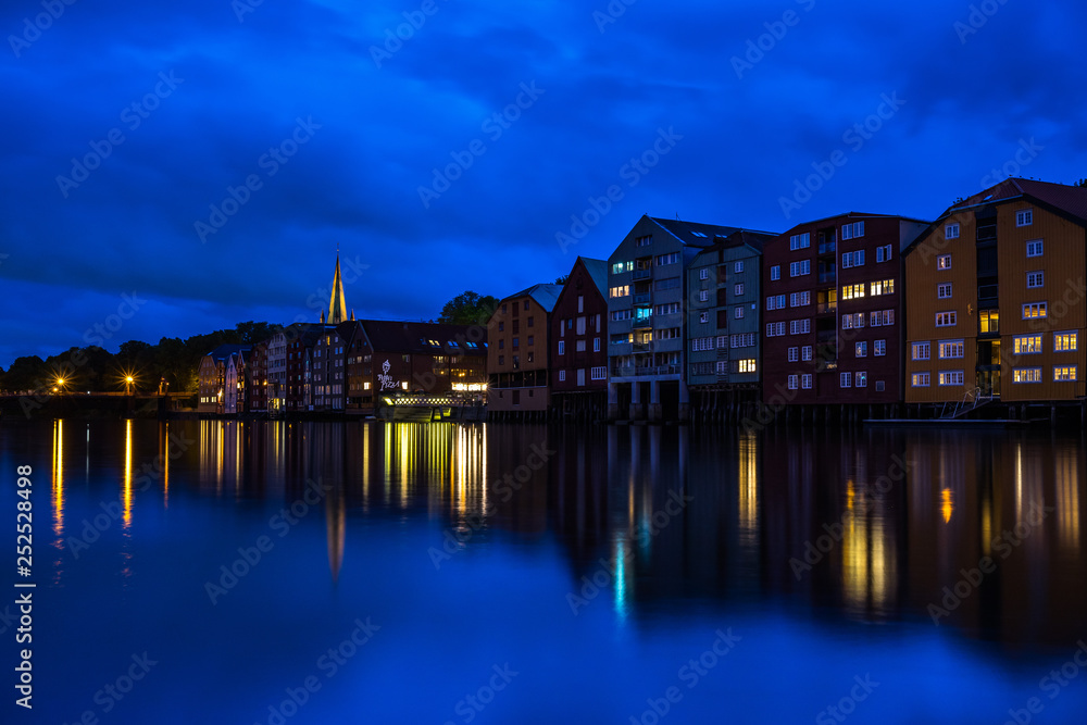 Trondheim cityscape at the blue hour with typical buildings along Nidelva River and the tower bell of Nidaros Cathedral in the background. Trondheim, Norway, August 2018