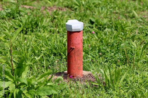 Industrial red partially rusted pipe covered with white metal cap on hard concrete foundation surrounded with uncut green grass and other small plants on warm sunny day