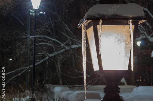 Lantern with an icicle glowing white light. Snow on the lantern, winter evening in the park.