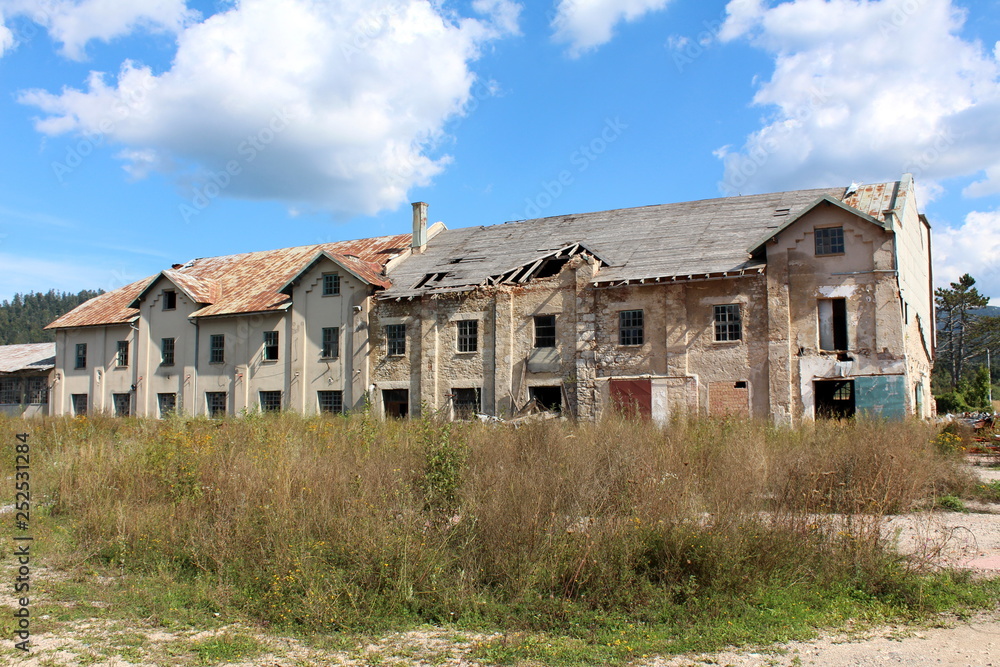 Large abandoned factory building with cracked dilapidated facade and destroyed roof surrounded with tall uncut grass and trees on cloudy blue sky background