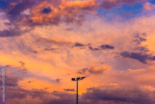 A cloudy orange sunset with the silhouette of a triple head sporting field light