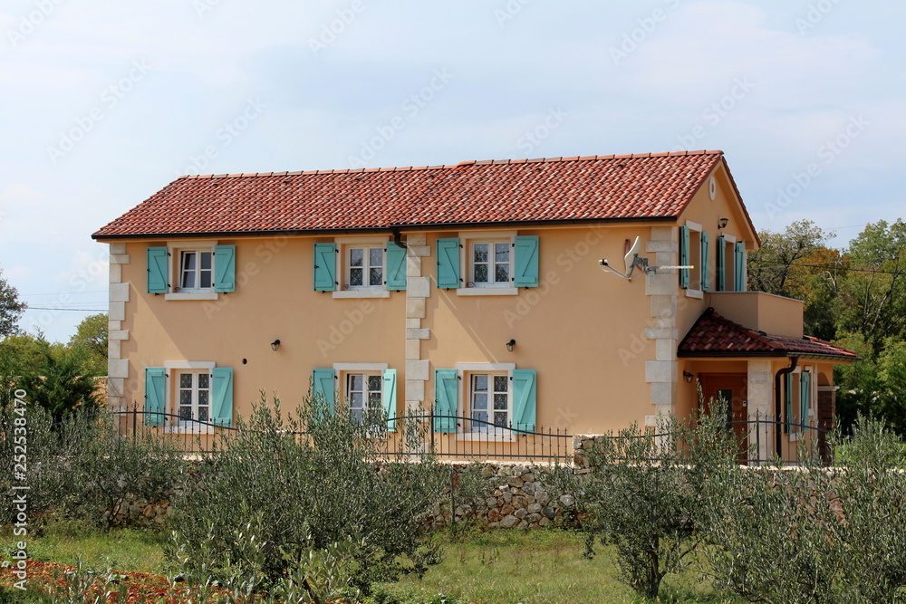 Newly built modern Mediterranean villa with new facade and open blue wooden window blinds with traditional stone wall and olive trees garden in front surrounded with dense trees and clear blue sky in 