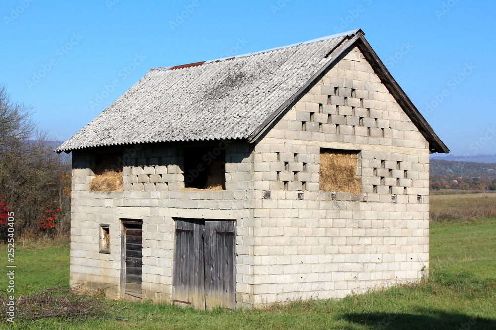 Outdoor concrete building blocks barn with dilapidated wooden doors full of hay surrounded with grass and trees on clear blue sky background Outdoor concrete building blocks barn with dilapidated wooden doors full of hay surrounded with grass and trees on clear blue sky background
