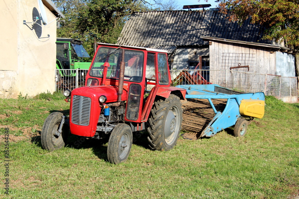 Obraz premium Red tractor parked next to family house with old agricultural equipment in background surrounded with barn and other tractor in background on warm sunny day