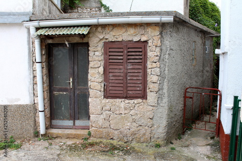 Stone and concrete small house with dilapidated cracked wooden doors and closed window blinds with new white metal gutter surrounded with concrete path on warm cloudy day