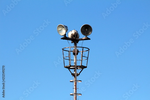 Tall metal pole with stairs leading to high; platform with three large electrical reflector lights on clear blue sky background