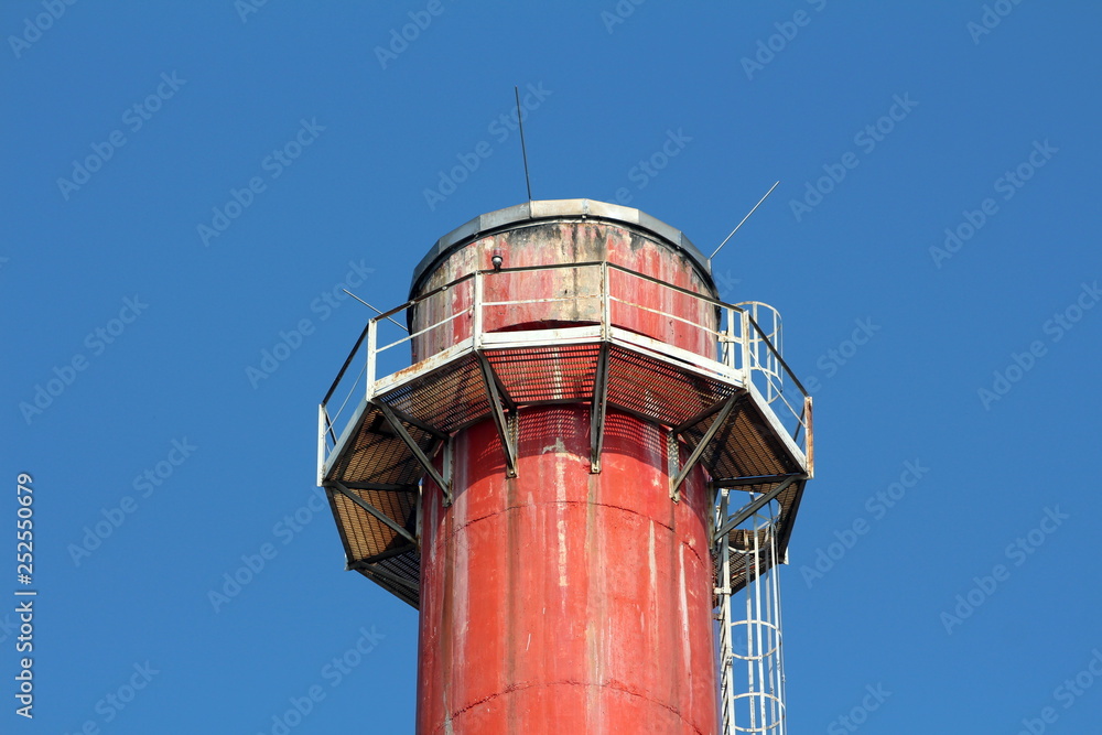 Top of old red tall industrial chimney surrounded with metal support platform and steps on clear blue sky background