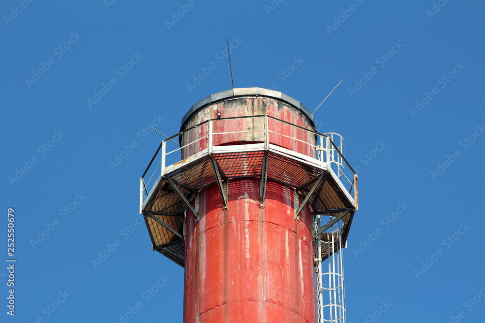 Top of old red tall industrial chimney surrounded with metal support platform and steps on clear blue sky background Top of old red tall industrial chimney surrounded with metal support platform and steps on clear blue sky background