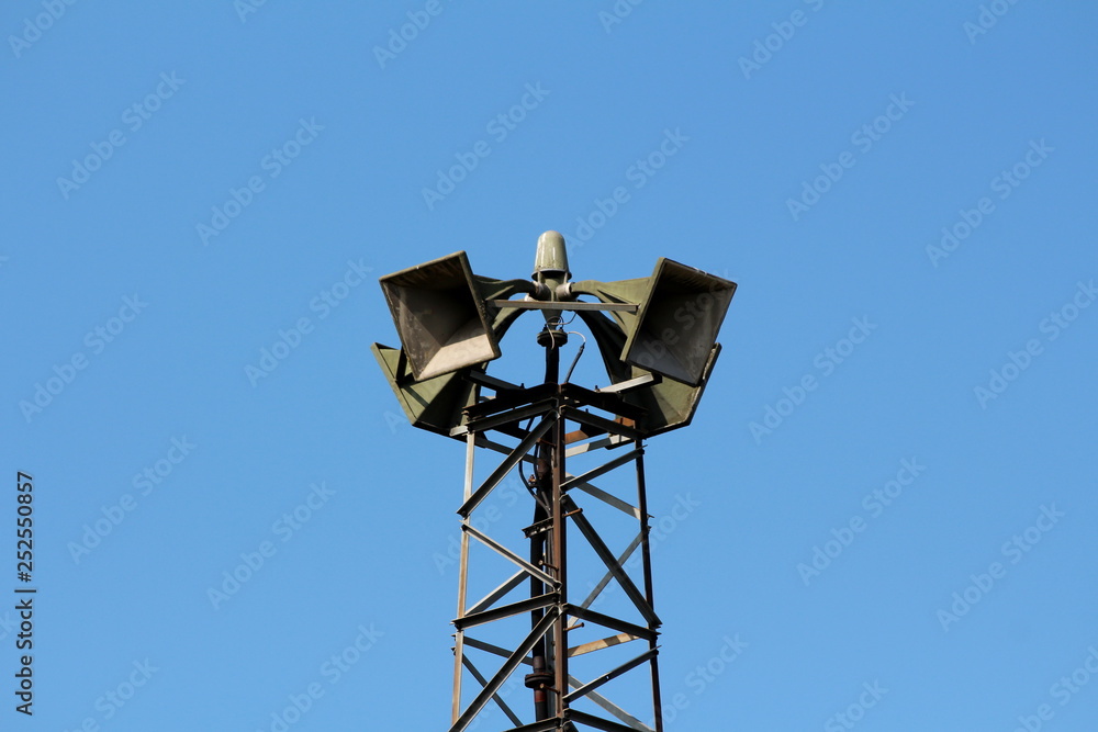 Top of tall metal structure holding four large public civil defence warning air sirens on clear blue sky background Top of tall metal structure holding four large public civil defence warning air sirens on clear blue sky background