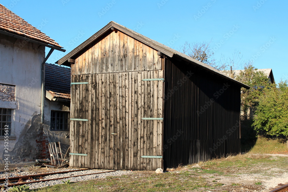 Wooden storage area locked with strong metal hinges and padlock with railroad tracks going inside for easier unloading surrounded with gravel and grass on clear blue sky background