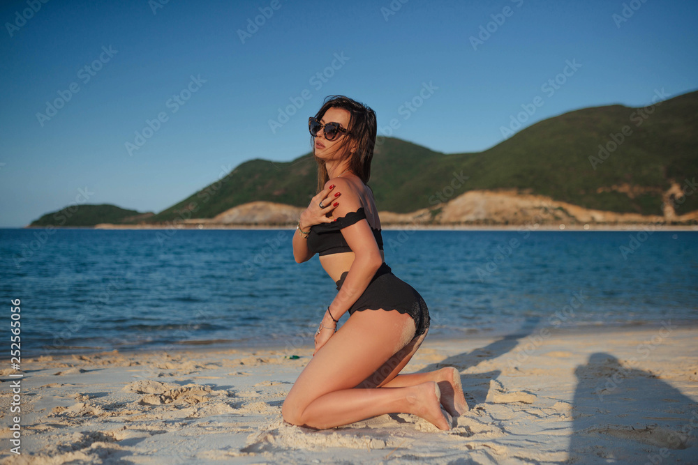  young beautiful girl, brunette, in a black bathing suit and sunglasses lies on the beach on white sand overlooking the sea. horizontal photo, large portrait 