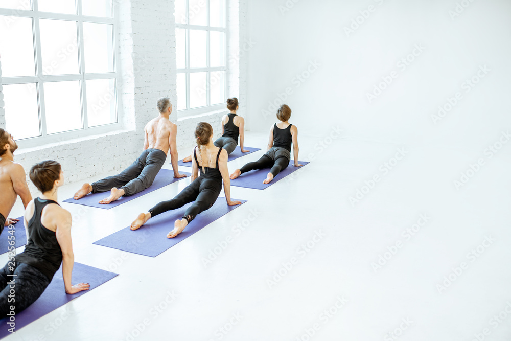 Young group of people practising yoga, keeping pose arranged together in a row in the white spacious studio. Wide view with copy space Young group of people practising yoga, keeping pose arranged together in a row in the white spacious studio. Wide view with copy space